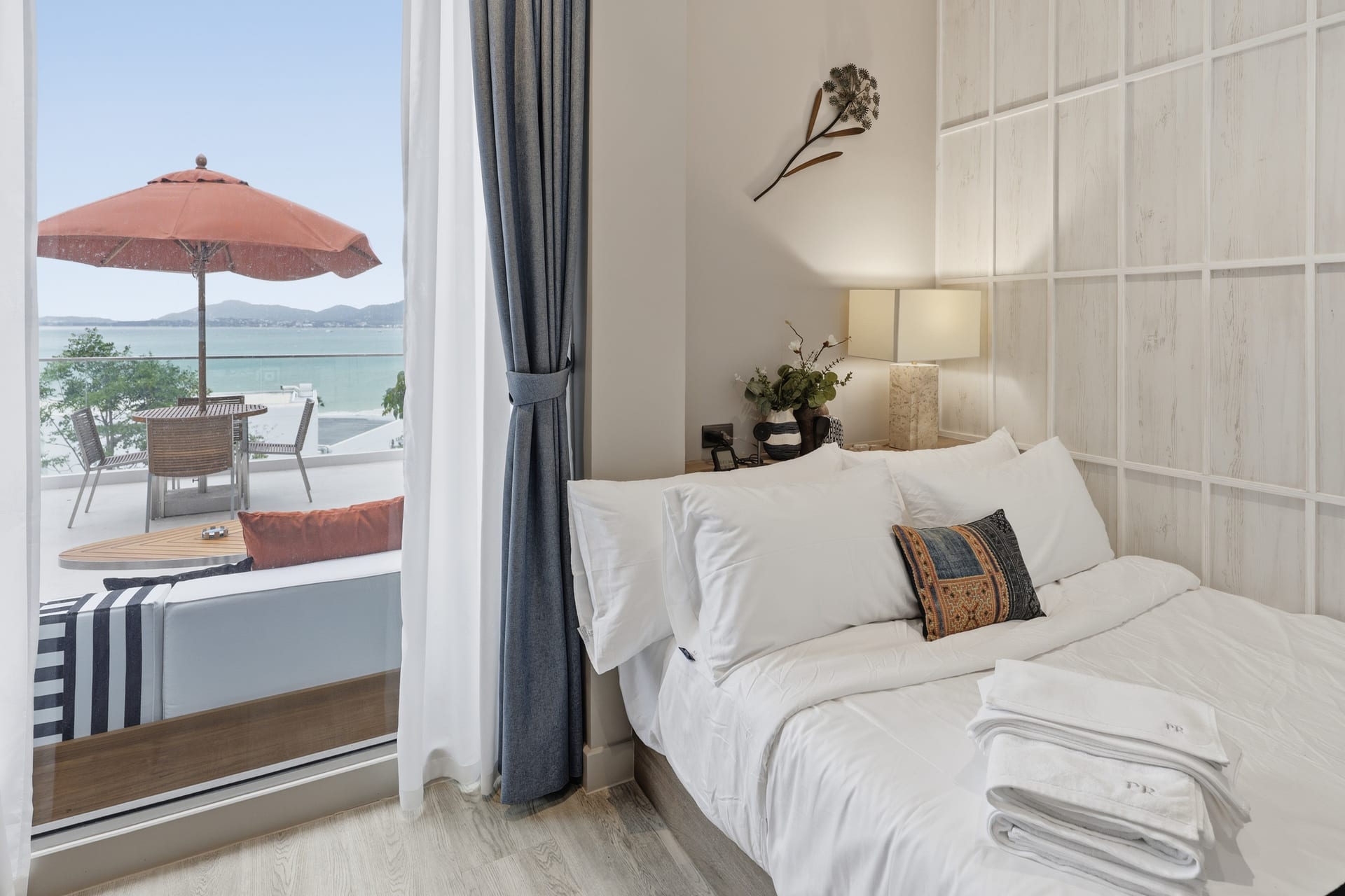 Bedroom with a queen bed in white linen and a patterned cushion, a bedside lamp, and sheer curtains framing a sliding door to a private balcony — a red parasol, table and chairs outside, and a daytime view across the bay to the mountains