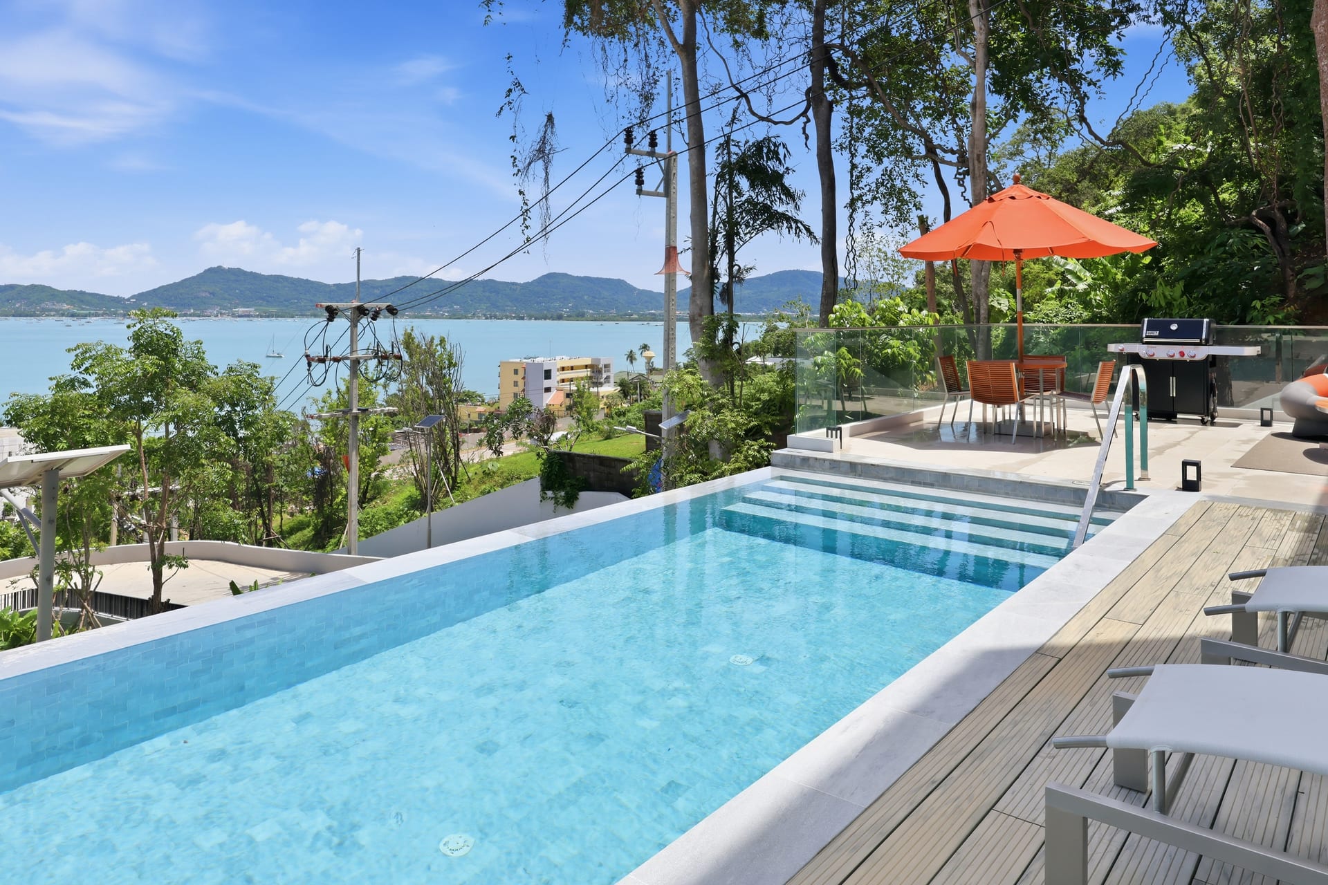 The private infinity pool on a clear sunny day — timber deck, an orange parasol and sun lounger, the infinity edge overlooking Ao Yon Bay with green headlands and the Phuket mountains under a bright blue sky