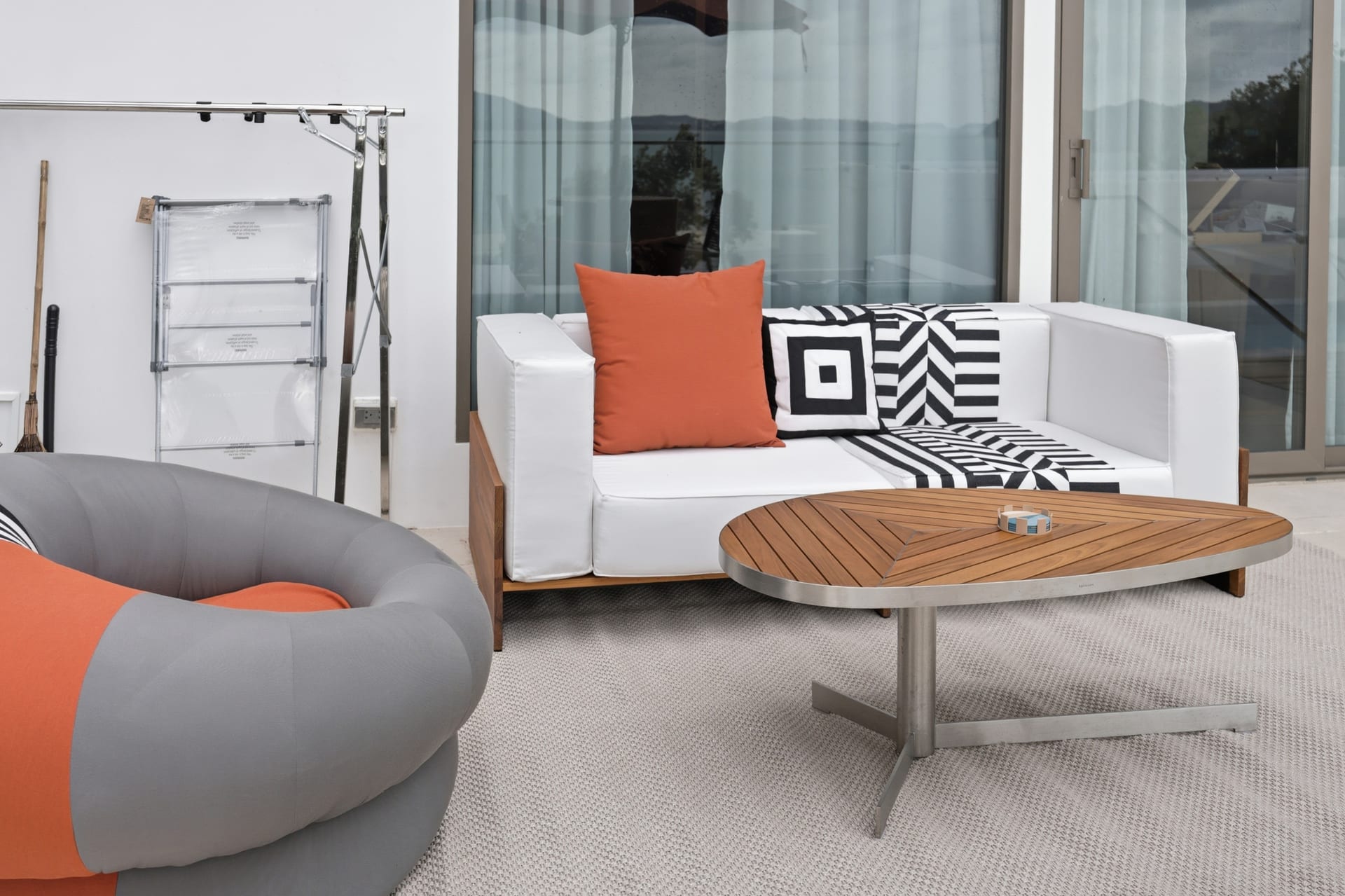 Terrace lounge seating — a white sofa with an orange cushion and a black-and-white geometric throw, a grey-and-orange rounded pod chair, and a round teak coffee table, with sheer curtains and a glimpse of the bay through the glass behind