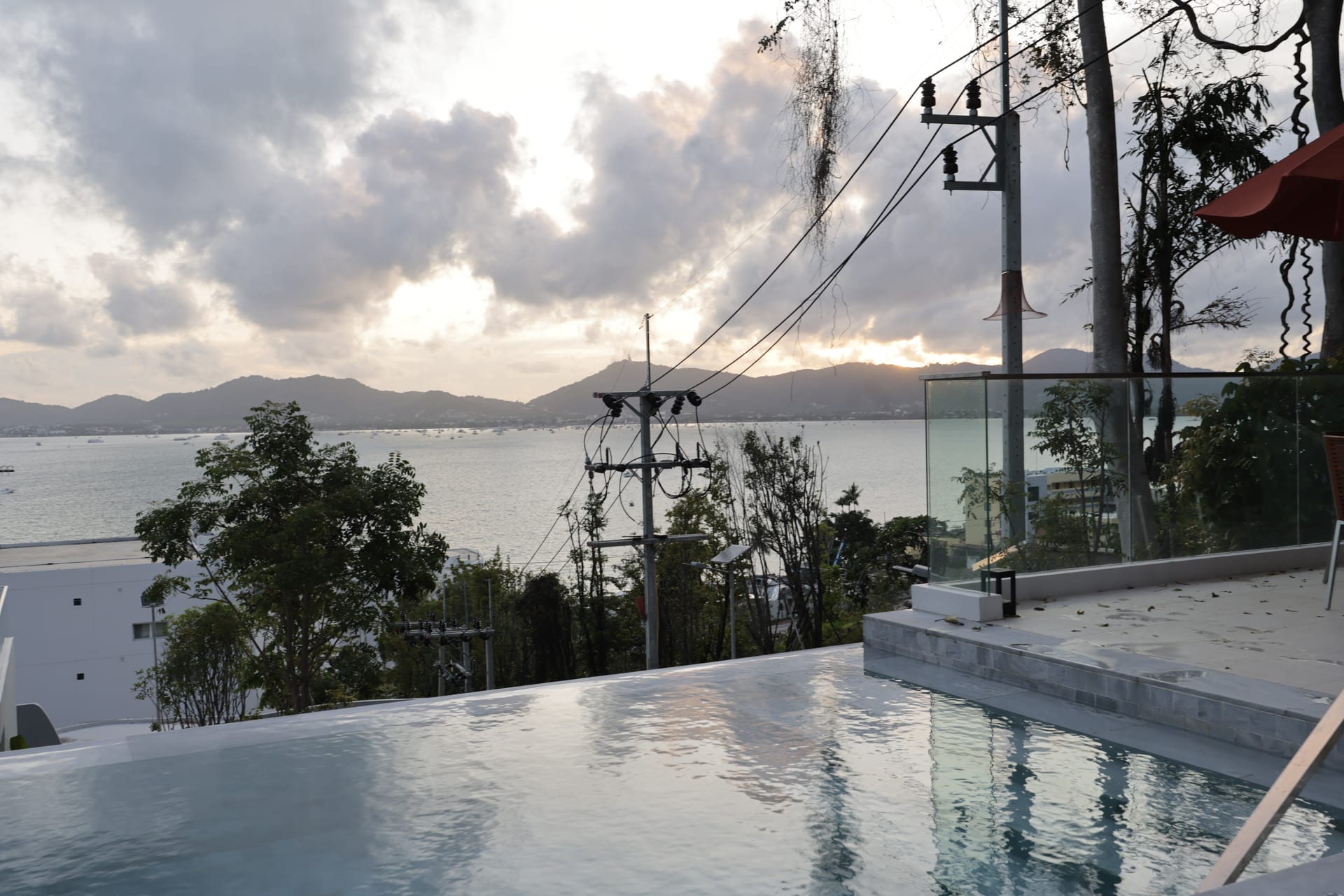 Infinity pool at dusk reflecting a soft pastel sky, with the bay, Chalong shoreline, and the Big Buddha hill silhouetted on the horizon — terrace parasol visible to the right