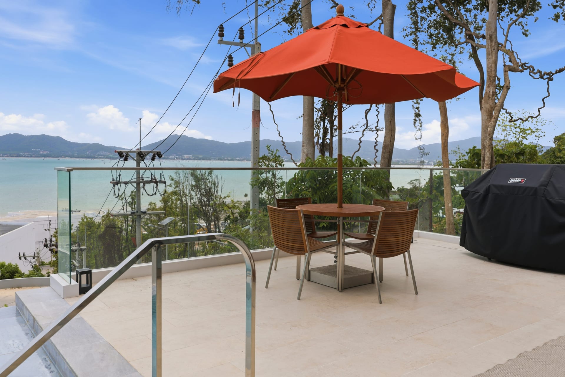 Al-fresco dining on the terrace under an orange parasol on a sunny day — the table set beside the glass railing with the bay and Cape Panwa hills behind