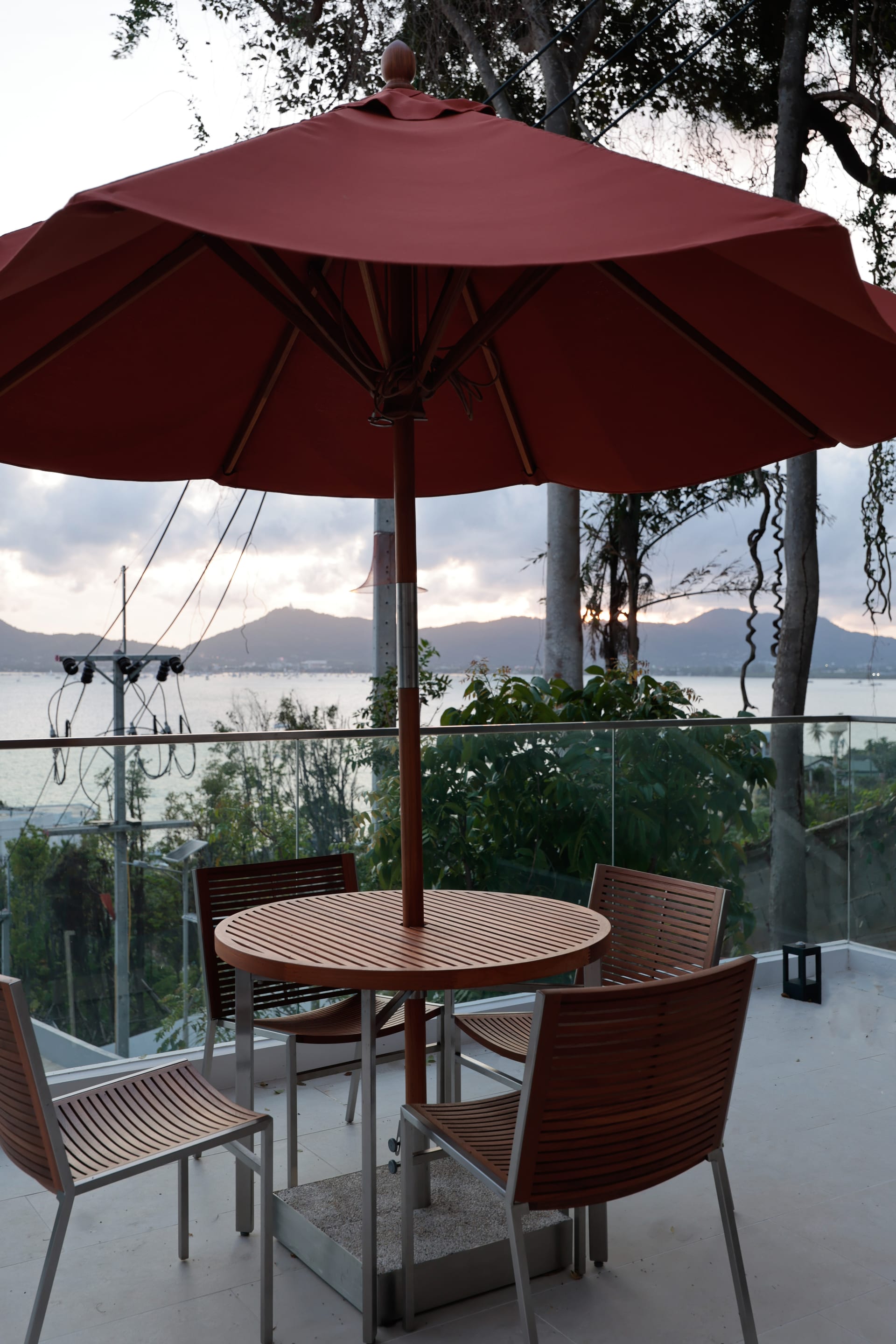 Outdoor dining setup on the terrace at dusk — wooden table with chairs under a deep-red parasol, glass railing opening to the bay and Cape Panwa hills behind