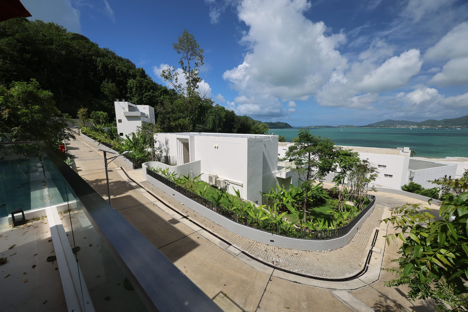 Alternate wide terrace view — glass railing in the foreground, hillside path on the left, neighbouring villa's planted garden in the middle ground, bay opening to the right under a bright sky