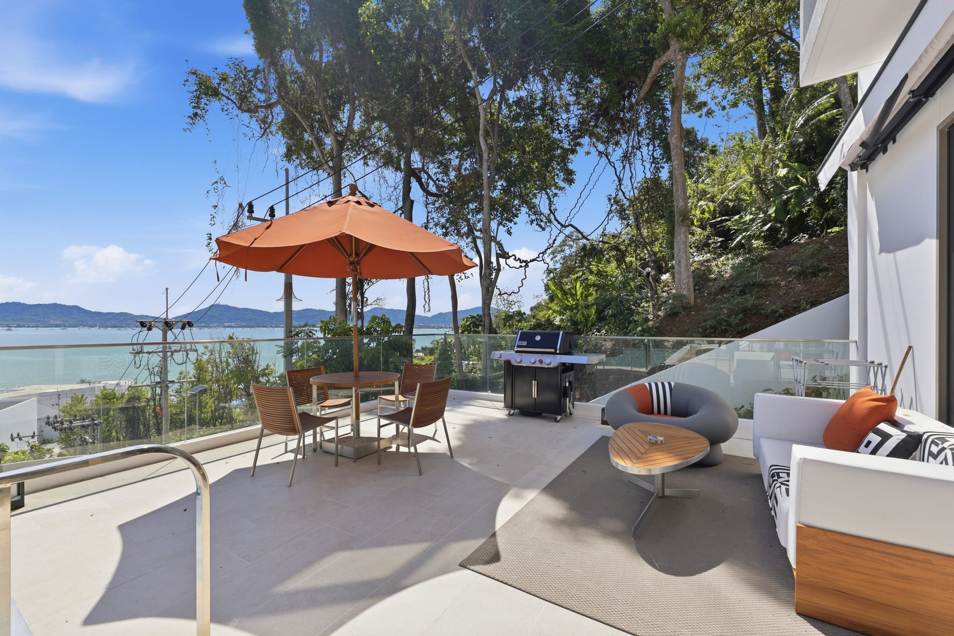 Terrace lounge seating under an orange parasol in bright sun, looking out over Ao Yon Bay and the Cape Panwa hills